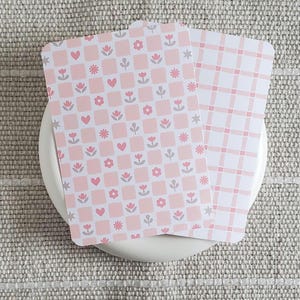 May include: Two decorative cards with rounded corners, featuring pink and white patterns. One card has a checkerboard design with hearts, flowers, and stars. The other card has a pink plaid pattern. The cards are on a white, round plate.