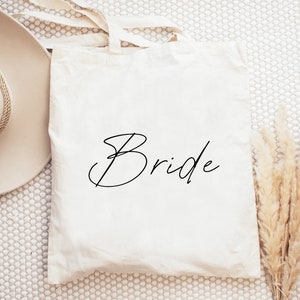 Puede incluir: Una bolsa de lona blanca con la palabra "Bride" escrita en cursiva negra.