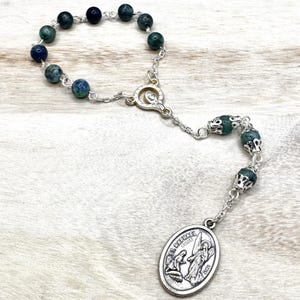 Puede incluir: Un rosario con cuentas verdes oscuras y azules, conectadas por una cadena plateada. El rosario presenta un colgante plateado con el texto "ST. RAPHAEL" y una imagen de un ángel. Las cuentas tienen aproximadamente 0,8 cm de diámetro.