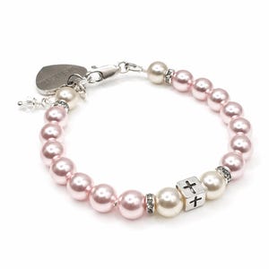 May include: A bracelet made of pink and white pearls, with a silver clasp and a heart-shaped charm. It includes a silver cube with a cross design and small crystal accents. The pearls are round and evenly spaced.
