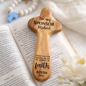 May include: A wooden cross with rounded edges, engraved with the words "For my SPONSOR Michael" and "THANK YOU FOR GUIDING ME ON MY JOURNEY IN faith AUSTIN 2026". The cross is resting on an open book, with a rosary and a blue cross in the background.