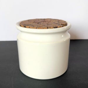 May include: A small, off-white ceramic jar with a rounded body and a cork lid. The jar has a smooth, glossy finish. The cork lid is circular and textured, providing a natural contrast to the jar's smooth surface. The jar is suitable for storing small items.
