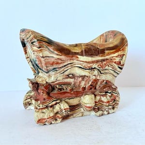 Puede incluir: Escultura abstracta artesanal con un diseño único en capas. La pieza presenta una mezcla de colores, incluyendo rojo, marrón, negro y crema, creando un efecto veteado. La escultura tiene una parte superior abierta y una superficie texturizada.