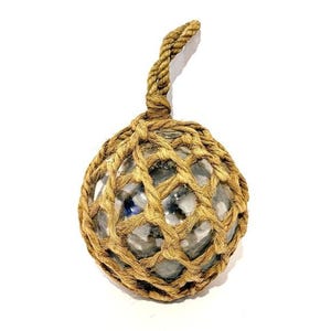 May include: A clear glass fishing float encased in a woven rope net. The float has a loop of rope at the top for hanging. The net and rope are a natural tan color. The float is a sphere shape.