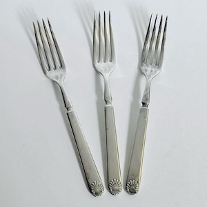 May include: Three silver forks with a shell design on the handle's end. The forks have four tines and a rectangular handle. The forks are arranged at different angles against a white background. The forks are likely vintage or antique.