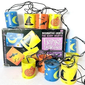 May include: A string of decorative Halloween lights with cylindrical shades. The shades are yellow, orange, and blue, featuring images of a witch's hat, bats, a jack-o'-lantern, a crescent moon, and stars. The box reads "Decorative Lights for Scary Nights!"