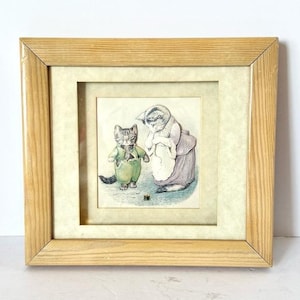 May include: Framed illustration featuring two anthropomorphic cats. One cat wears a green outfit, the other a purple dress and apron. The artwork is set against a cream-colored background, framed in light wood.