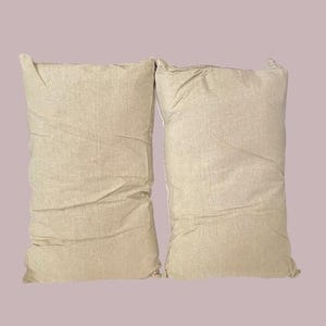 May include: Two beige pillowcases made of a soft, textured fabric. The pillowcases are lying flat on a light purple background.