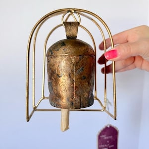 May include: A vintage-style bell with a textured, bronze-colored body and a wooden clapper. The bell is suspended within a gold-colored metal frame. A small tag is attached to the frame. The bell is held by a person.