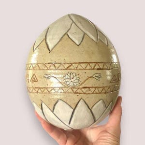 May include: A ceramic egg-shaped vase with a beige glaze and white and brown painted designs. The vase features a geometric pattern, a stylized flower, and a series of overlapping leaf shapes.