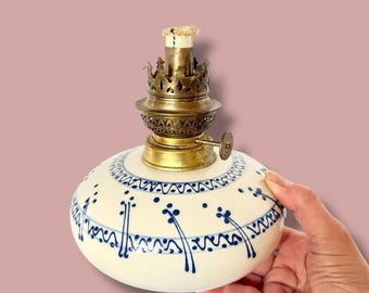 May include: A vintage oil lamp with a white ceramic base adorned with blue floral patterns. The lamp features a brass burner and wick, with a decorative crown-shaped top. The lamp is round and held by a hand.