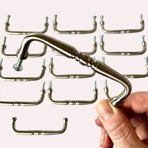 May include: A collection of brushed nickel-finished cabinet pulls. One pull is held up with a screw, showcasing its design. The pulls have a curved handle and are designed for easy grip. The hardware is ideal for kitchen or bathroom cabinets.