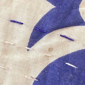 May include: Close-up of a white fabric with blue stitching and a blue crescent moon shape. The fabric has a rough texture and is slightly wrinkled.