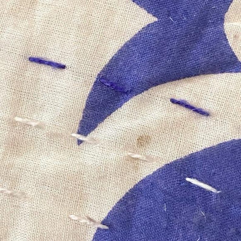 May include: Close-up of a white fabric with blue stitching and a blue crescent moon shape. The fabric has a rough texture and is slightly wrinkled.