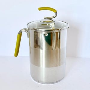 May include: A stainless steel coffee press with a glass lid and a yellow handle and lid grip. The cylindrical body has a polished finish. The handle is on the side, and the lid grip is arched. The coffee press is designed for brewing coffee.