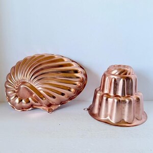 May include: Two copper-colored molds. One is a seashell shape, the other is a tiered cake shape. Both have detailed designs and are made of shiny metal. These vintage molds are ideal for baking and kitchen decor.