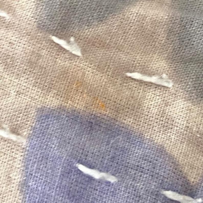 May include: Close-up of a beige and light blue fabric with white stitching. The fabric has a subtle texture and a light brown stain.