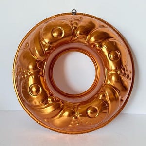 May include: A vintage copper bundt cake pan with a decorative fruit design. The pan has a ring shape with a central hole and a hanging loop. The copper has a warm, reflective finish.