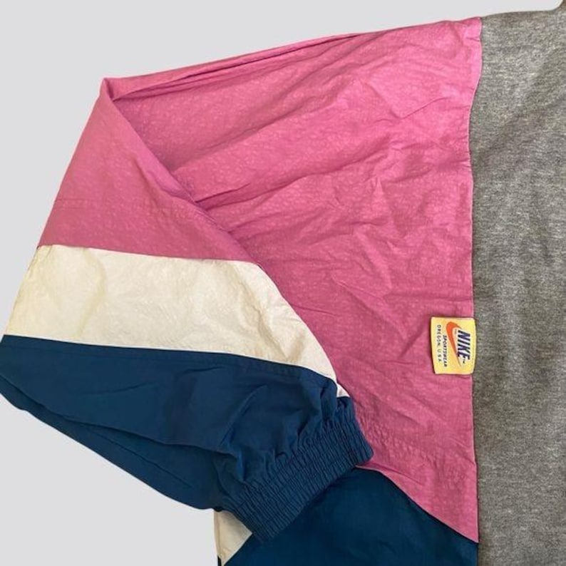 May include: A blue, white, and pink windbreaker jacket with a Nike logo patch. The jacket has a zipper closure and a drawstring hood.