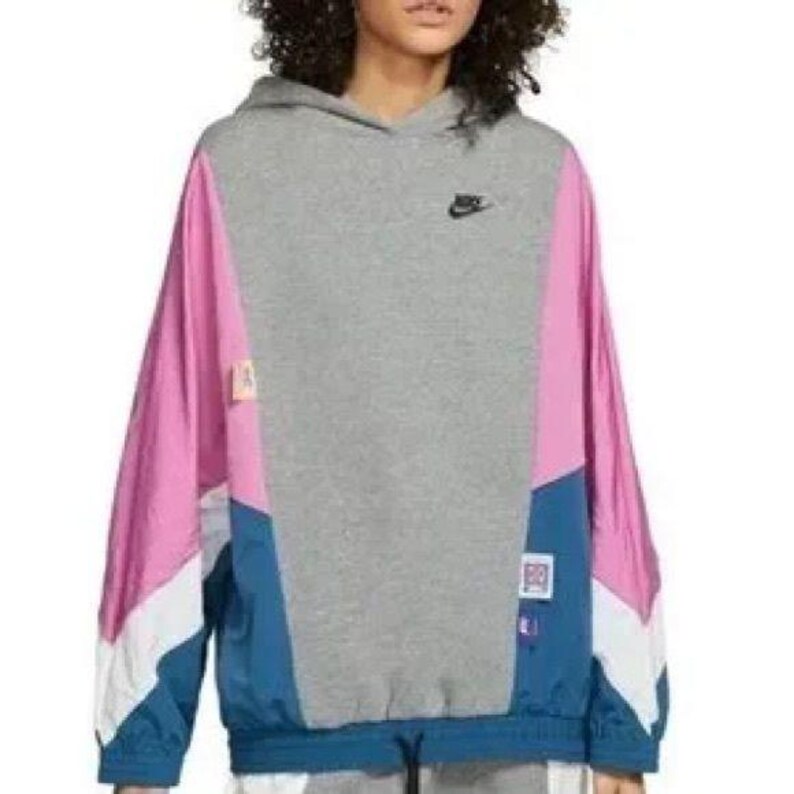 May include: A gray hooded sweatshirt with pink, blue, and white color-blocked panels. The sweatshirt has a Nike logo on the left chest and a small patch on the left sleeve.
