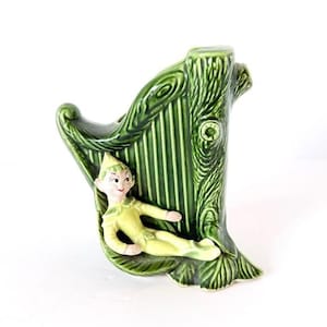 May include: A green ceramic vase shaped like a tree trunk and harp, with a small elf figure resting on the harp. The vase has a glossy finish and is painted in shades of green and yellow. The elf is wearing a yellow outfit.