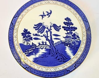 May include: Round blue and white decorative plate with a Willow pattern design. The plate features a central landscape scene with a bridge, trees, and buildings, framed by a patterned border. The plate has a gold trim.