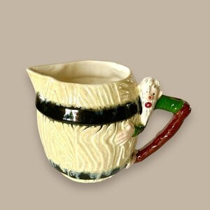 May include: A vintage ceramic pitcher with a cream-colored, textured barrel design and a black band. The handle is a brown, green, and white figure. The pitcher has a spout for pouring.