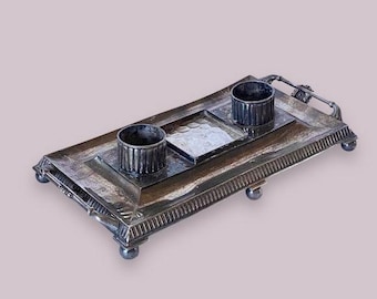 May include: A silver inkwell with two inkwells and a central tray. The inkwell has a decorative border and sits on four feet.