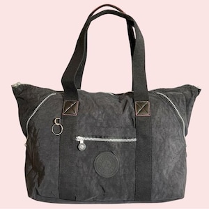 May include: A black tote bag with two handles and a shoulder strap. The bag has a textured appearance, multiple zippered compartments, and a circular logo patch. The zippers and hardware are silver-toned.