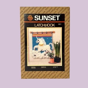 May include: A Sunset latchhook kit featuring a whimsical unicorn design. The kit includes a framed image of a white unicorn on a cloud, with the text "MAGICAL UNICORN" and "LATCHHOOK" on the packaging. Finished size is 18" x 18".