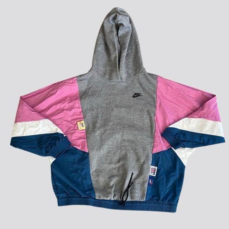 May include: A gray hooded sweatshirt with pink, white, and blue color-blocked panels. The sweatshirt has a Nike logo on the front and a drawstring at the bottom.