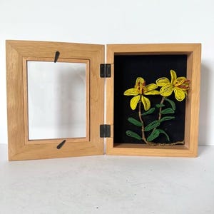 May include: A wooden framed piece of art with a hinged door. The artwork features yellow beaded flowers with green leaves on a black background. The frame is light brown and the hinges are black.