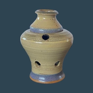 May include: A handmade ceramic garlic keeper in a beige and blue color scheme. The garlic keeper has a bulbous body with three holes and a lid with a single hole. The pottery has a rustic, textured appearance.