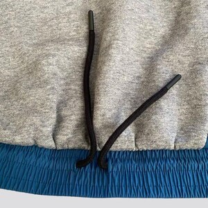 May include: Close-up of a gray sweatshirt with a blue elastic waistband and black drawstring. The drawstring is tied in a knot at the center.
