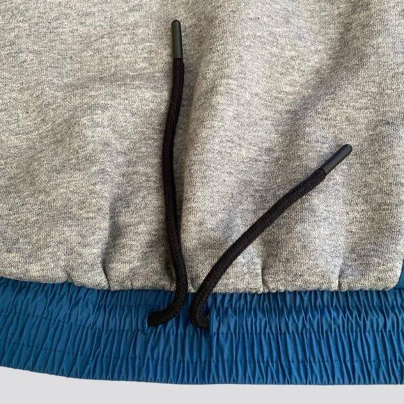 May include: Close-up of a gray sweatshirt with a blue elastic waistband and black drawstring. The drawstring is tied in a knot at the center.