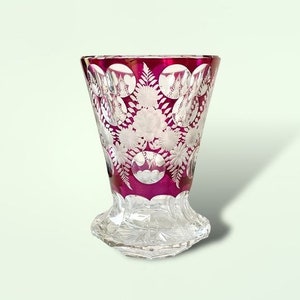 May include: A ruby red glass vase with a cut glass design of white flowers and leaves. The vase has a flared top and a clear glass base.