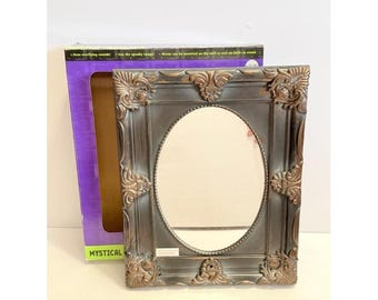 May include: Ornate, rectangular mirror with an oval centre. The frame is dark grey with bronze-coloured accents. The mirror is next to a purple box with the word "MYSTICAL" printed on it.