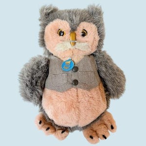 May include: A plush owl toy with gray and peach-colored fur. The owl wears a gray herringbone vest with two black buttons and a blue tag. The owl has yellow eyes and a metal nose piece.