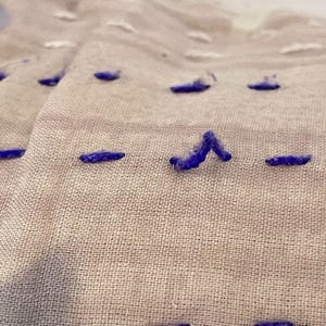 May include: Close-up of a beige fabric with purple stitching in a straight line and a V-shape.