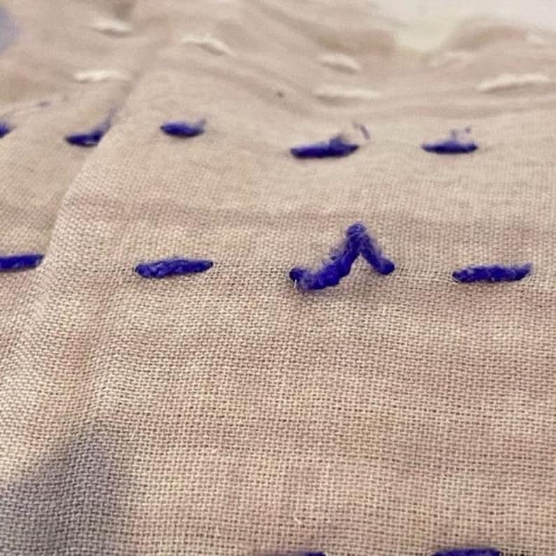 May include: Close-up of a beige fabric with purple stitching in a straight line and a V-shape.