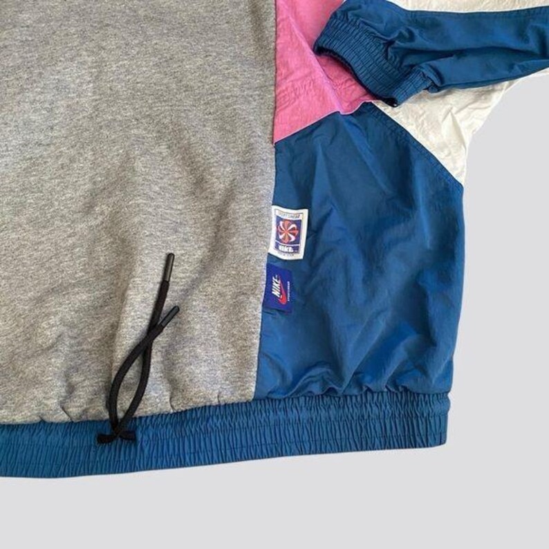 May include: A blue, pink, white, and gray color block jacket with a drawstring at the bottom. The jacket has a Nike logo and a small logo with a star on it.
