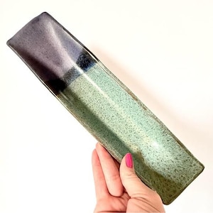 May include: A rectangular ceramic serving dish with a gradient color scheme. The dish transitions from a dark purple at one end to a speckled teal green. The dish is held by a hand with pink nail polish against a white background.