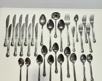 May include: A collection of vintage silverware, including knives, spoons, forks, and a ladle, arranged on a white surface. The silverware is silver-toned with ornate handle detailing, reflecting light. The arrangement suggests a formal dining setting.