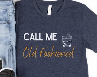 May include: A heather navy blue t-shirt with the text "CALL ME Old Fashioned" in orange and white. The shirt features a graphic of a glass of old fashioned with a lemon twist.