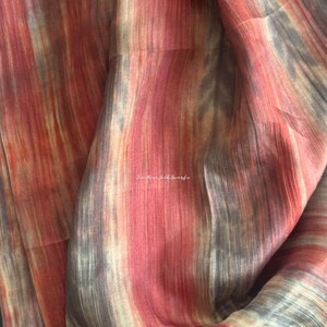 May include: A lightweight fabric featuring a vertical, abstract pattern in shades of rust orange, muted grey, and cream. The material has a subtle, semi-sheer texture with visible fibres. The text "JinRose Silk Scarfs" is printed in white in the centre.