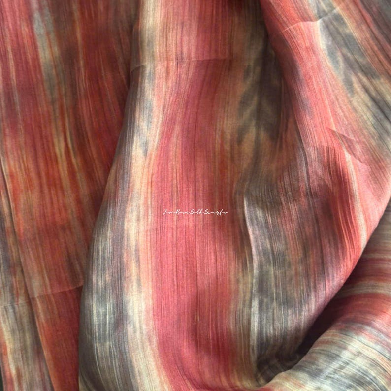 May include: A lightweight fabric featuring a vertical, abstract pattern in shades of rust orange, muted grey, and cream. The material has a subtle, semi-sheer texture with visible fibres. The text "JinRose Silk Scarfs" is printed in white in the centre.