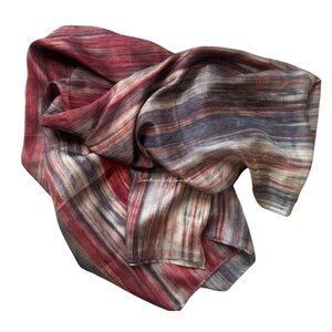 May include: A lightweight scarf featuring a multi-coloured striped pattern in shades of deep red, charcoal grey, and cream. The fabric displays a subtle, flowing texture with horizontal lines throughout. The text "Kim Rose Silk Scarfs" is visible.