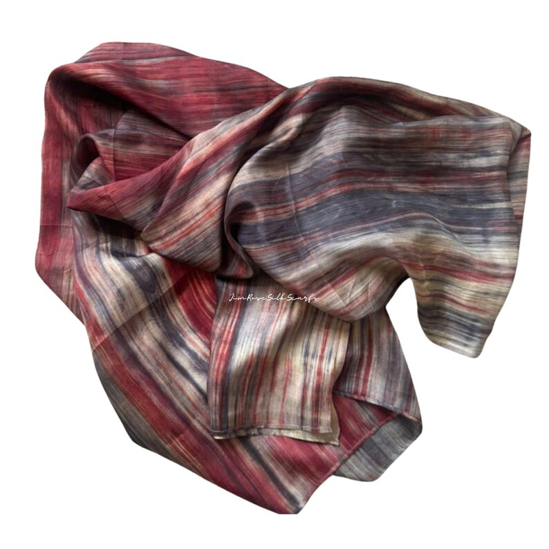 May include: A lightweight scarf featuring a multi-coloured striped pattern in shades of deep red, charcoal grey, and cream. The fabric displays a subtle, flowing texture with horizontal lines throughout. The text "Kim Rose Silk Scarfs" is visible.