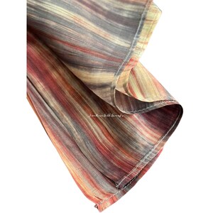 May include: A lightweight fabric scarf featuring a vertical striped pattern in shades of rust, tan, and charcoal grey. The material has a subtle sheen and visible stitching along the hem. The text Jim Rose Silk Scarf is printed on the fabric.