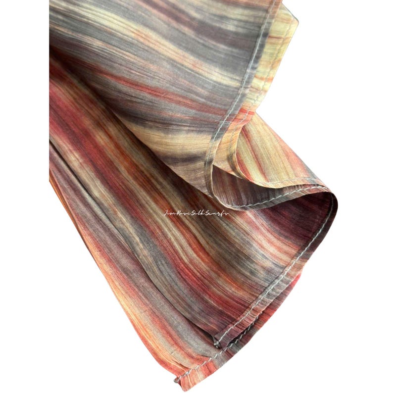 May include: A lightweight fabric scarf featuring a vertical striped pattern in shades of rust, tan, and charcoal grey. The material has a subtle sheen and visible stitching along the hem. The text Jim Rose Silk Scarf is printed on the fabric.